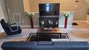 Una sala de estar con un sofá negro, una mesa de centro negra, un televisor de pantalla plana y dos jarrones rojos con flores blancas.