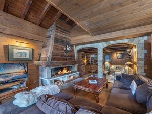 A cozy log cabin interior with a stone fireplace, leather furniture, and a stone archway leading to another room.