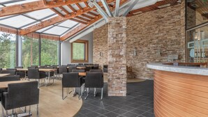 A modern indoor space with a high ceiling, exposed wooden beams, and a stone wall. Tables and chairs are arranged for dining or meetings.