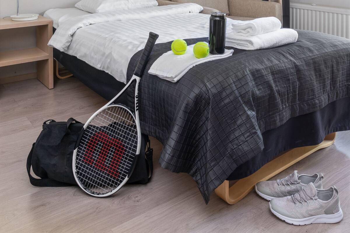 Un lit bien fait, avec une couette à carreaux gris, une raquette de tennis et des ballons de sport posés sur le sol à côté.
