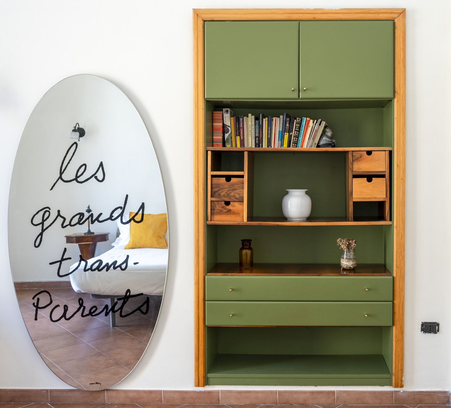 A green bookshelf with books and decorative items, a white wall with a mirror displaying text, and a bed with a yellow pillow.