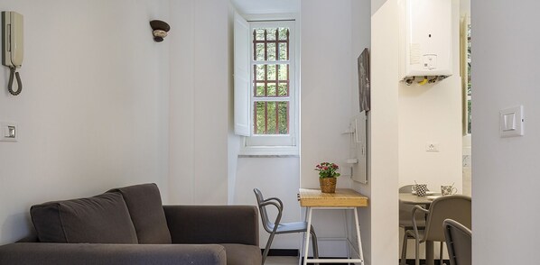 Ett kompakt vardagsrum med en soffa, ett litet bord med en växt, en matplats med stolar och ett fönster med utsikt över grönområden.