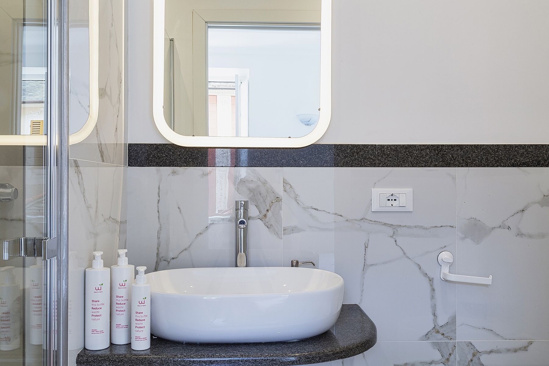 A bathroom with a white sink, marble walls, a glass shower enclosure, and a mirror.