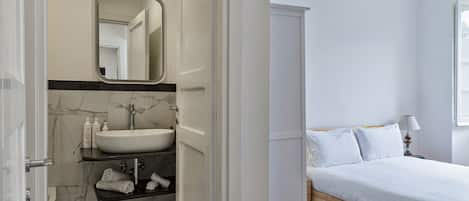 A bathroom with a marble sink, a mirror, and a shower. Adjacent is a bedroom with a bed and a nightstand.