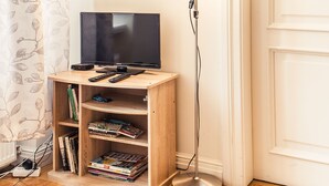 Habitación de esquina con un mueble bajo de madera para televisor, una pantalla plana, una lámpara de pie y una estantería con libros y revistas.