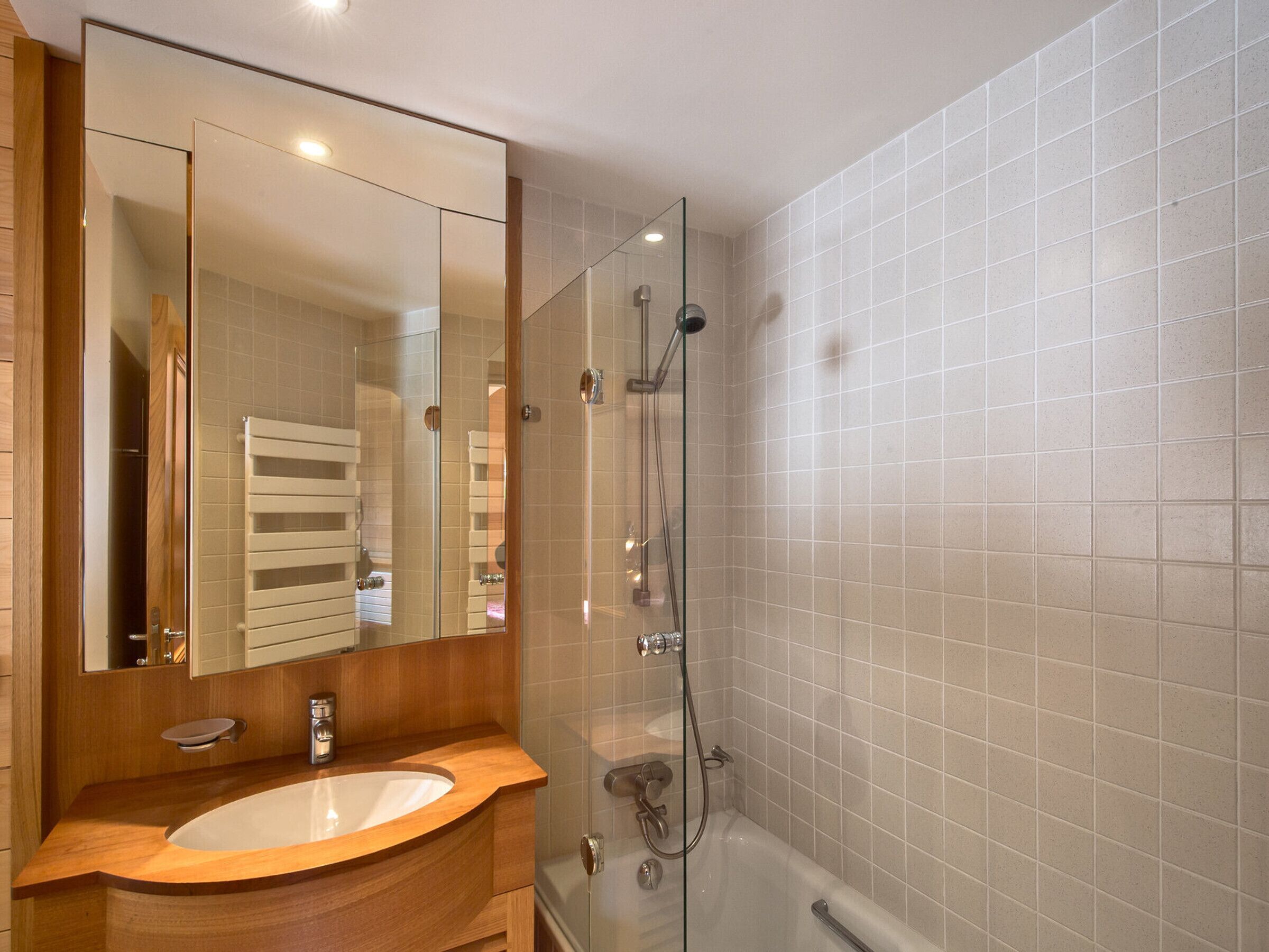 Un baño moderno con tocador de madera, un espejo grande, cabina de ducha de vidrio y paredes de azulejos blancos.