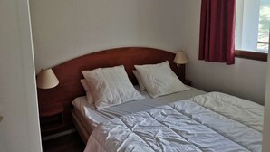 A hotel room with a wooden headboard, two white pillows, a white bedspread, a nightstand with a lamp, and a window with a red curtain. - Apartment with mountain view - 4 guests (La Mongie)