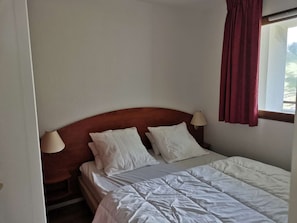 Quarto de hotel com cabeceira de madeira, duas almofadas brancas, colcha branca, mesa de cabeceira com abajur e janela com cortina vermelha.