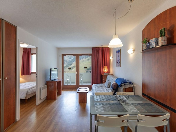 Quarto compacto com cama, mesa de jantar com cadeiras, uma mesinha com abajur e vista para o exterior por uma porta de vidro.