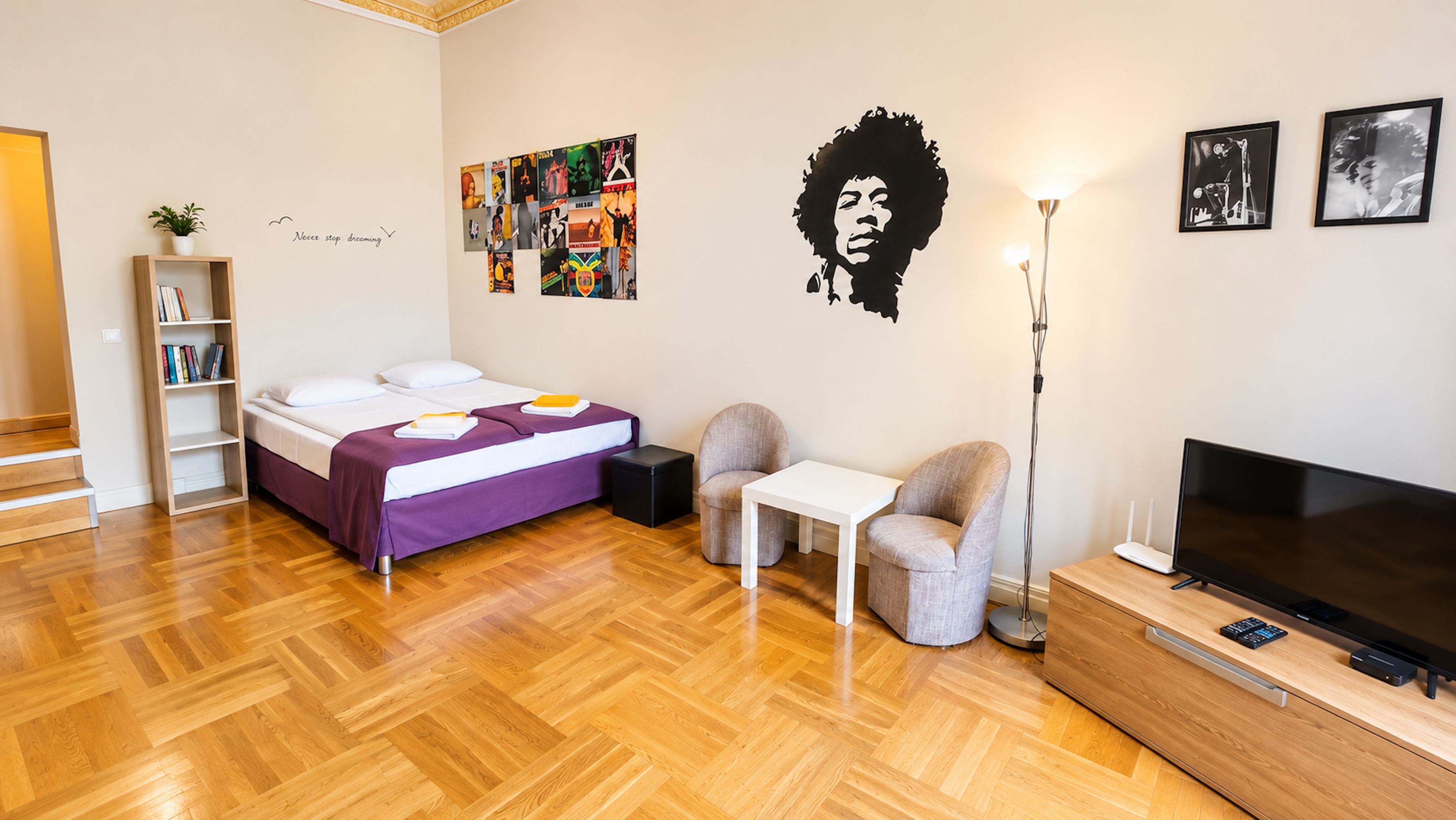 Ein modernes Schlafzimmer mit Bett, Schreibtisch, Stuhl, Fernseher und einer Wand mit verschiedenen Postern und einem motivierenden Zitat.