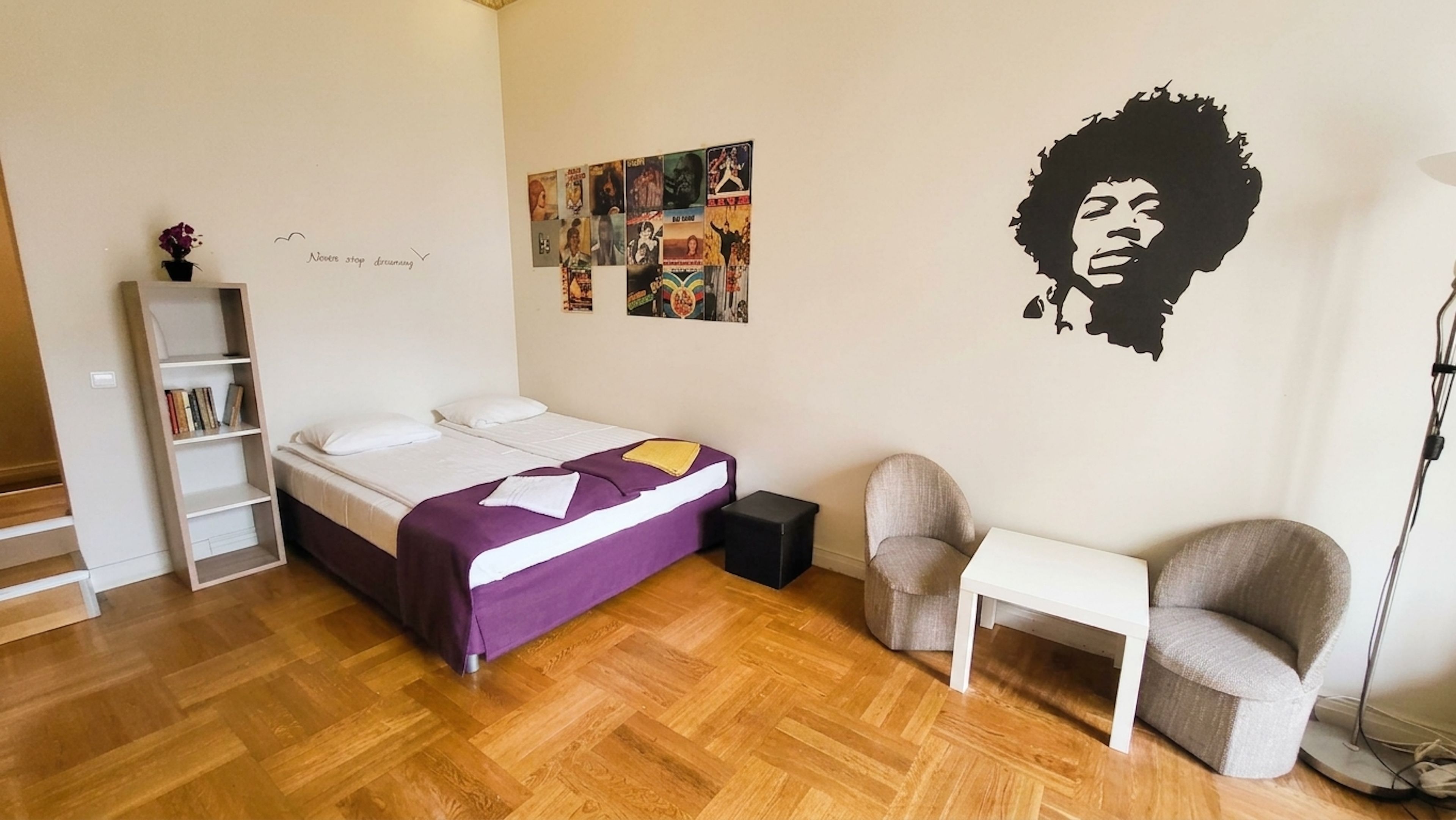 Une chambre à coucher avec un lit, une chaise, un bureau, une bibliothèque et un mur orné d’une affiche.