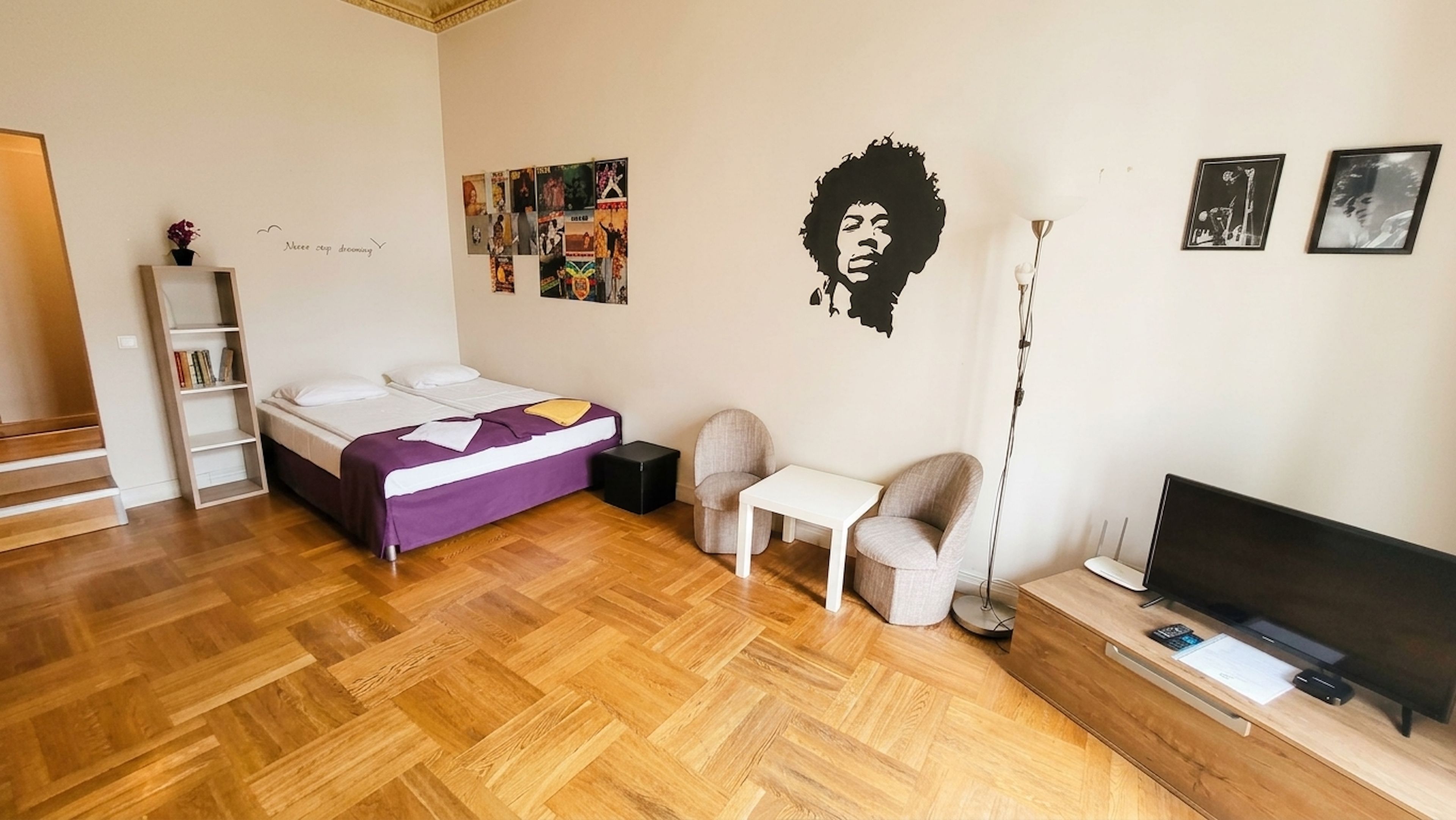 A bedroom with a bed, a desk, a chair, a TV, and a wall with various pictures and a mural.