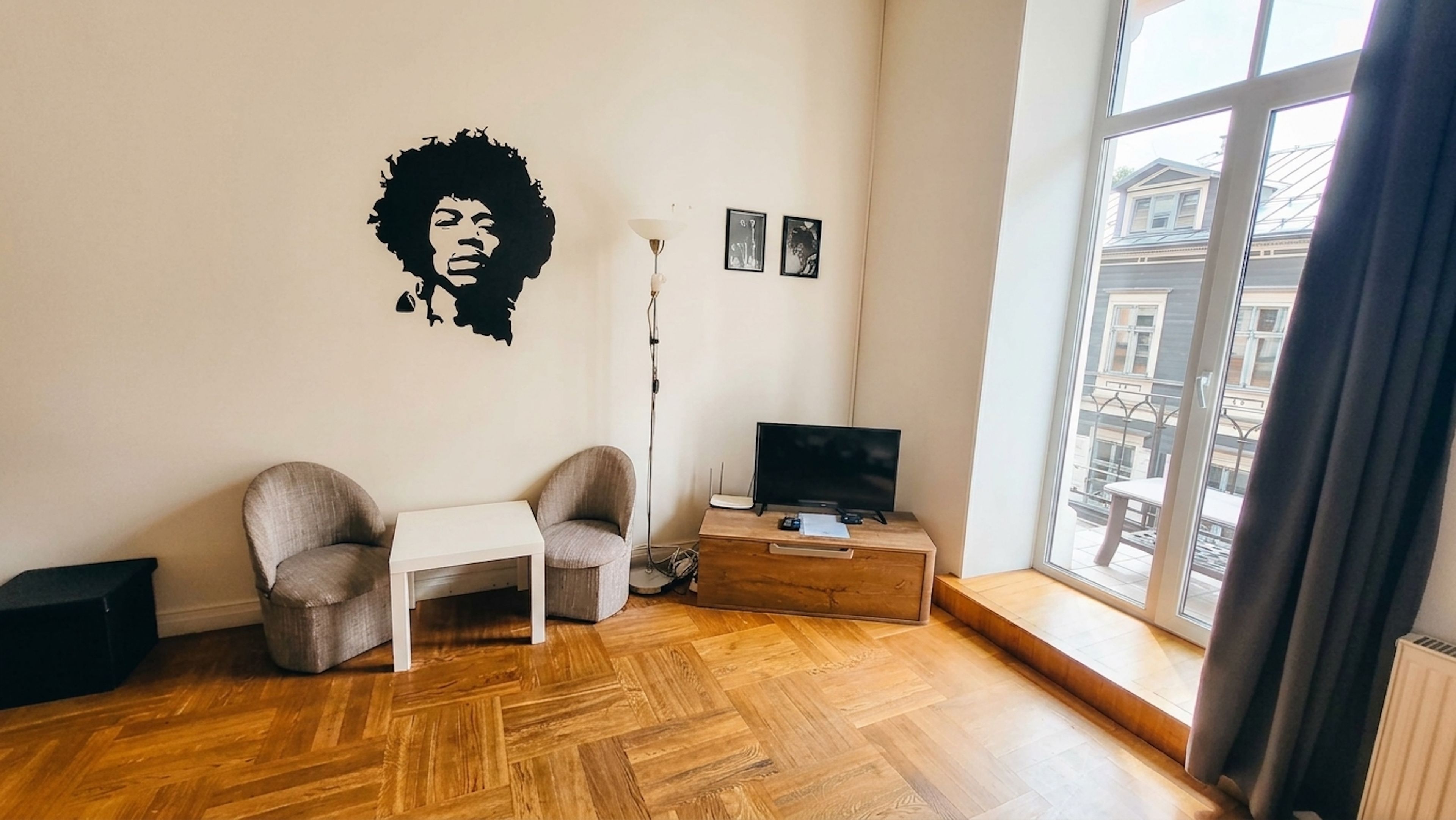 Une pièce avec un plancher en bois, un téléviseur posé sur un meuble en bois, deux fauteuils, une petite table blanche et une grande fenêtre donnant sur des immeubles.