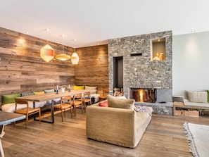 Uma sala de estar moderna com lareira de pedra, paredes de madeira e uma área de jantar com uma mesa comprida e cadeiras.