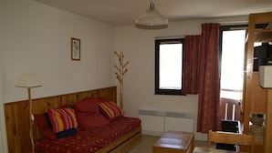 A room with a wooden bed, a small table, a lamp, a window with curtains, and a radiator.