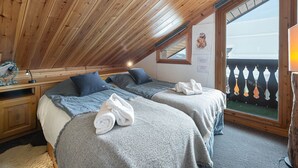 A cozy attic bedroom with wooden ceiling beams, two beds with grey bedding, a small desk, and a window overlooking a balcony.