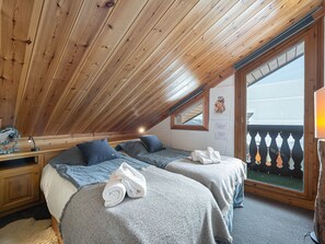 Um quarto de sótão aconchegante com vigas de madeira no teto, duas camas com roupa de cama cinza, uma pequena mesa de escritório e uma janela com vista para uma varanda.