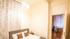 Una cama bien hecha con cabecera gris, una mesita de noche con lámpara, y una puerta que da a un pasillo con suelo de madera.