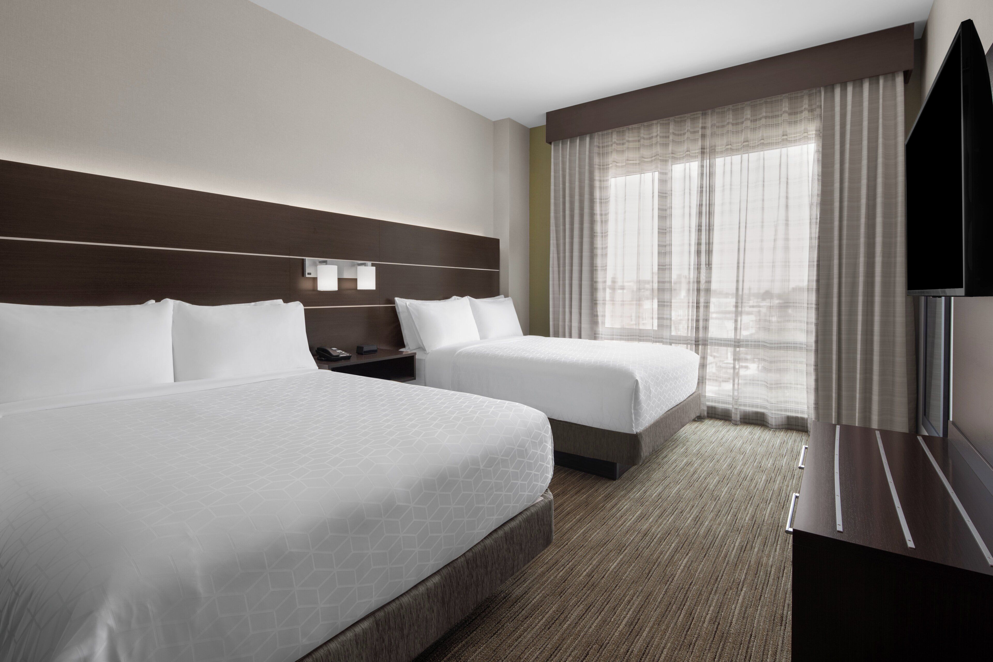 A hotel room with two beds, a flat-screen TV, a nightstand, and a large window with curtains.