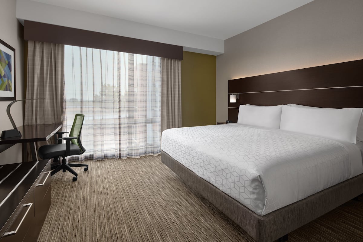 Ein Hotelzimmer mit einem großen Bett, einem Schreibtisch mit Stuhl, einem Fenster mit Vorhängen und einem Kopfteil mit integrierter Beleuchtung.