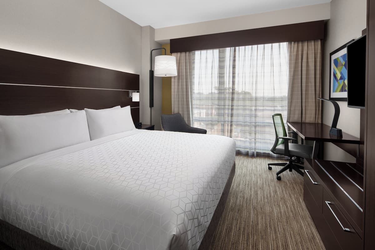 Ein Hotelzimmer mit einem großen Bett, einem Schreibtisch mit Stuhl, einem Fernseher und einem Fenster mit Vorhängen.