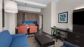 Una habitación de hotel moderna con un sofá azul, una pequeña mesa de comedor con sillas naranjas, una kitchenette, un escritorio con silla y un televisor de pantalla plana montado en la pared.