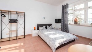 Ein Schlafzimmer mit Bett, Fernseher, Fenster mit Vorhängen, Nachttisch und Kleiderschrank mit darin befindlichen Kleidungsstücken.
