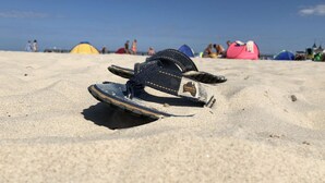 Sandalen auf sandigem Strand, im Hintergrund Zelte und Personen.