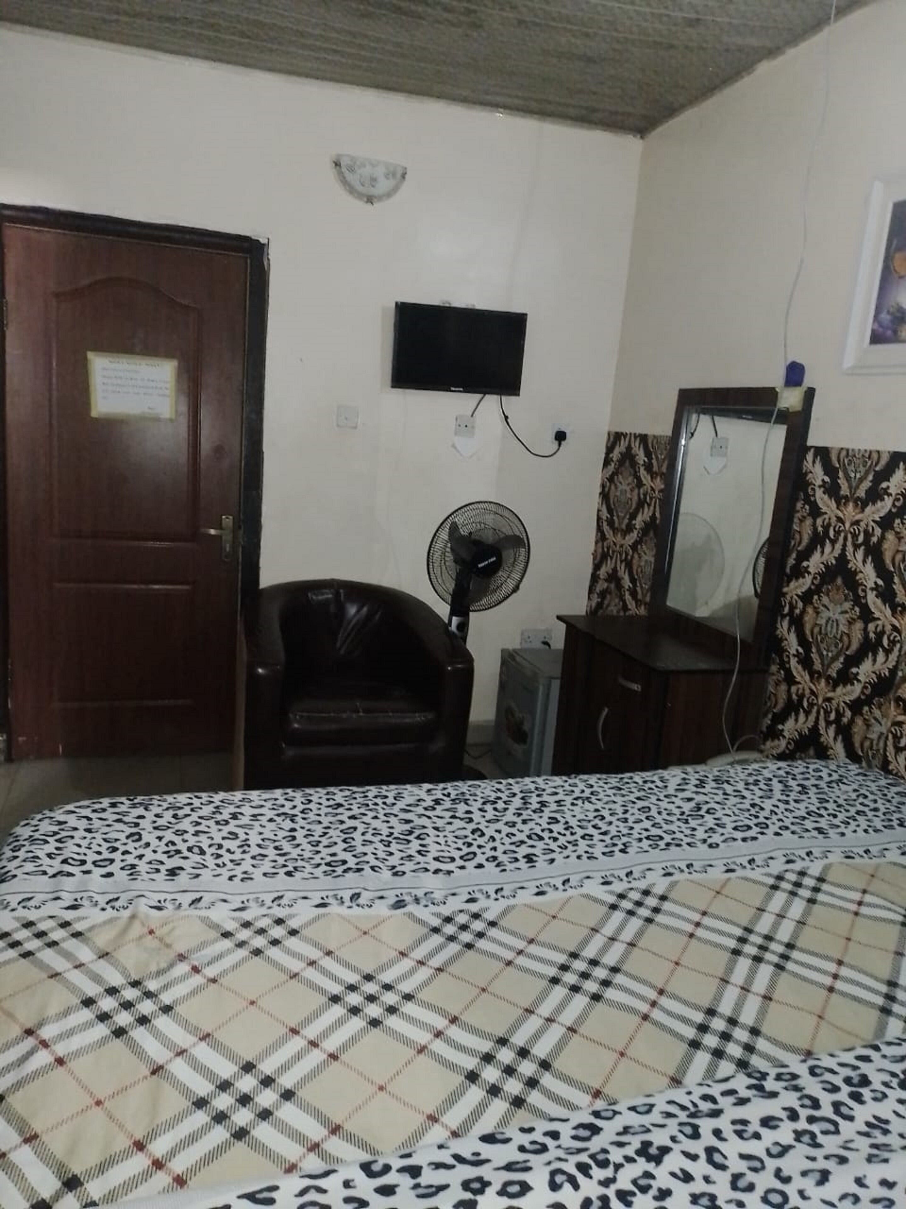 A bedroom with a bed, a desk, a chair, a fan, a television, and a wardrobe.