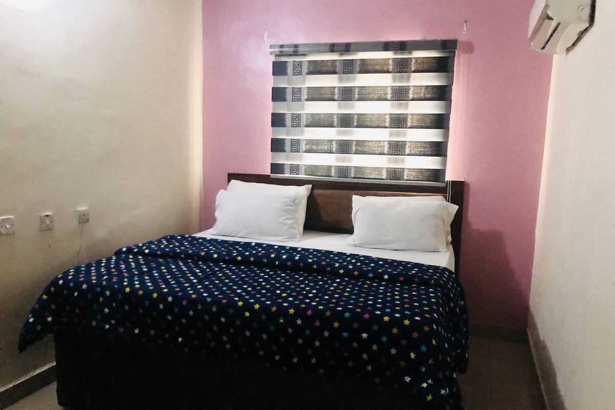 A bedroom with a bed, a window with patterned curtains, and a pink wall.