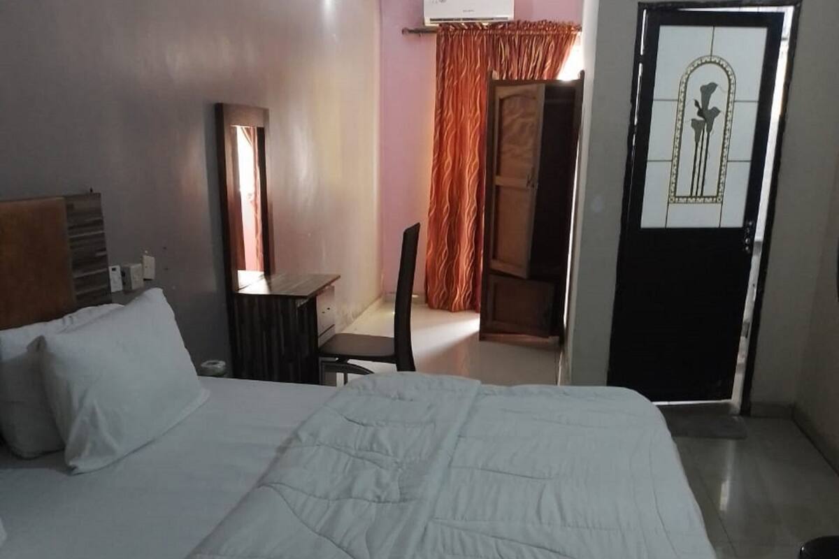 A hotel room with a large bed, a desk, a chair, an air conditioning unit, and a door with a mirror.