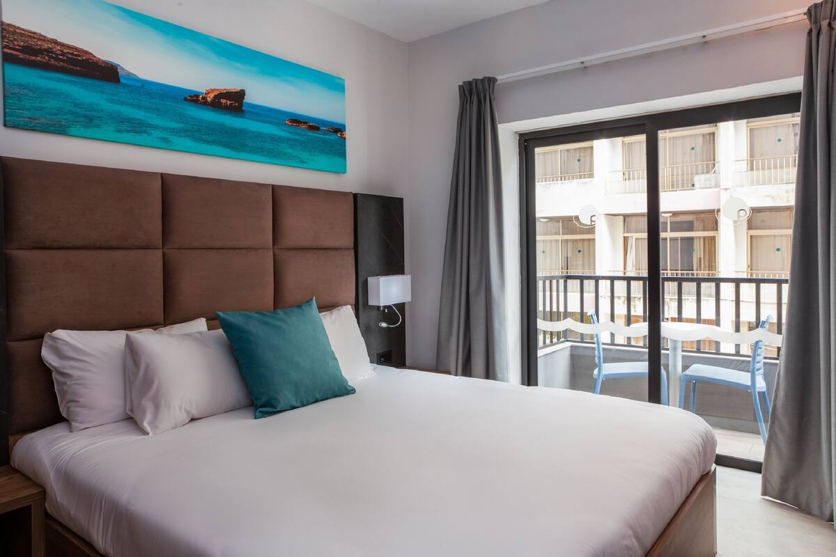 Ein Hotelzimmer mit einem großen Bett, einem Gemälde einer Küstenlandschaft, einem Balkon mit Stühlen und Blick auf ein Gebäude.