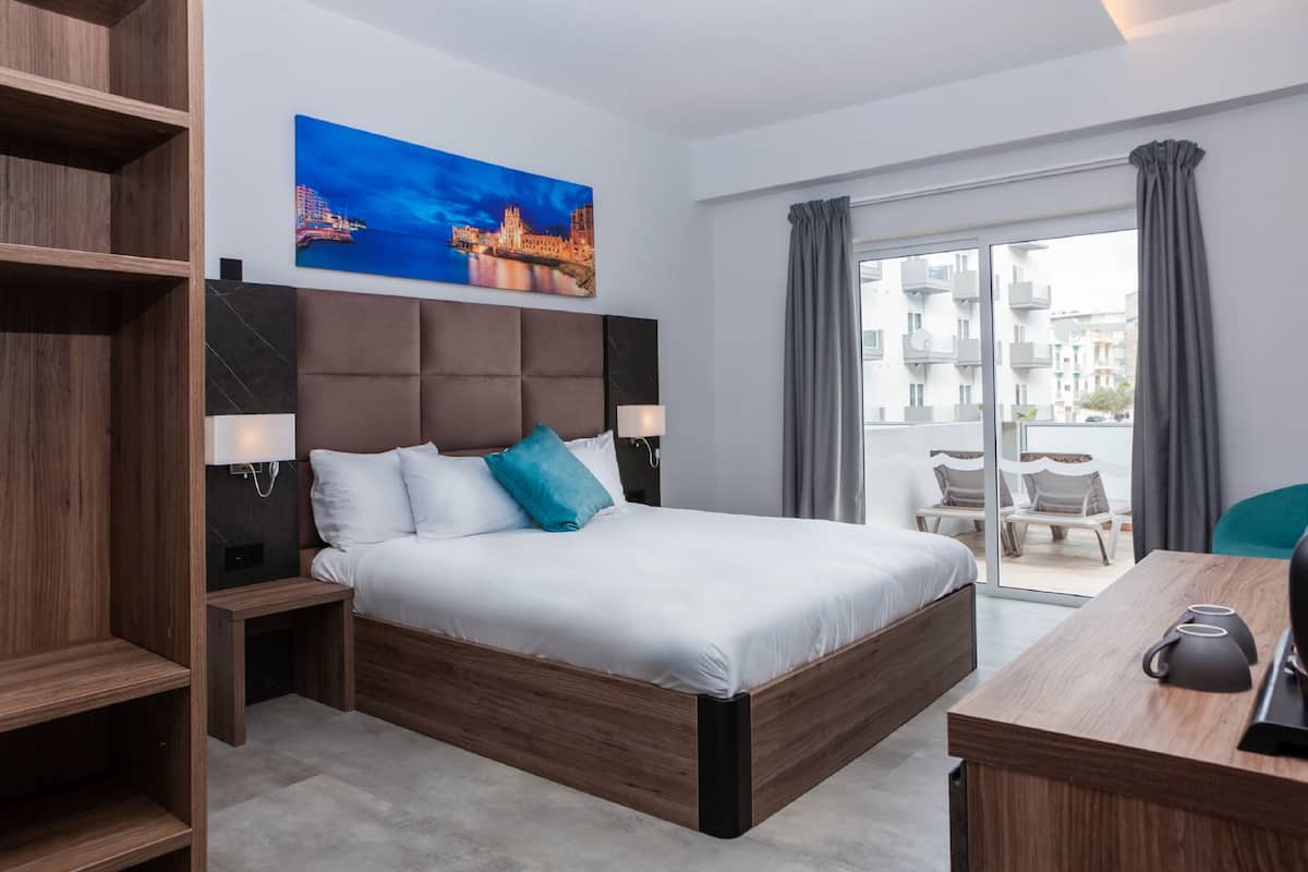 Ein modernes Hotelzimmer mit einem großen Bett, einem Holzkopfende und einem Stadtbildgemälde. Ein Schreibtisch mit Stuhl und Blick auf die Stadt durch große Fenster.