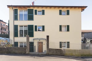 Un edificio de dos pisos con fachada color crema, contraventanas verdes y una entrada protegida por una reja.