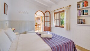 A hotel room with a bed, a bookshelf, a window with curtains, and a door leading to another room. - Finca “Ses Begudes” with Garden, Terrace, Pool, Air Conditioning & Wi-Fi (Fornalutx)