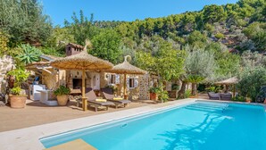 A swimming pool with lounge chairs, a stone building with a thatched roof, and surrounding greenery. - Finca “Ses Begudes” with Garden, Terrace, Pool, Air Conditioning & Wi-Fi (Fornalutx)