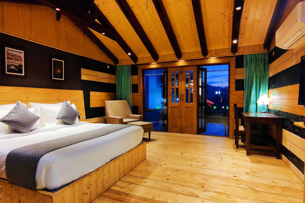 A wooden-floored bedroom with a large bed, two chairs, a desk, and a sliding glass door leading to a balcony.