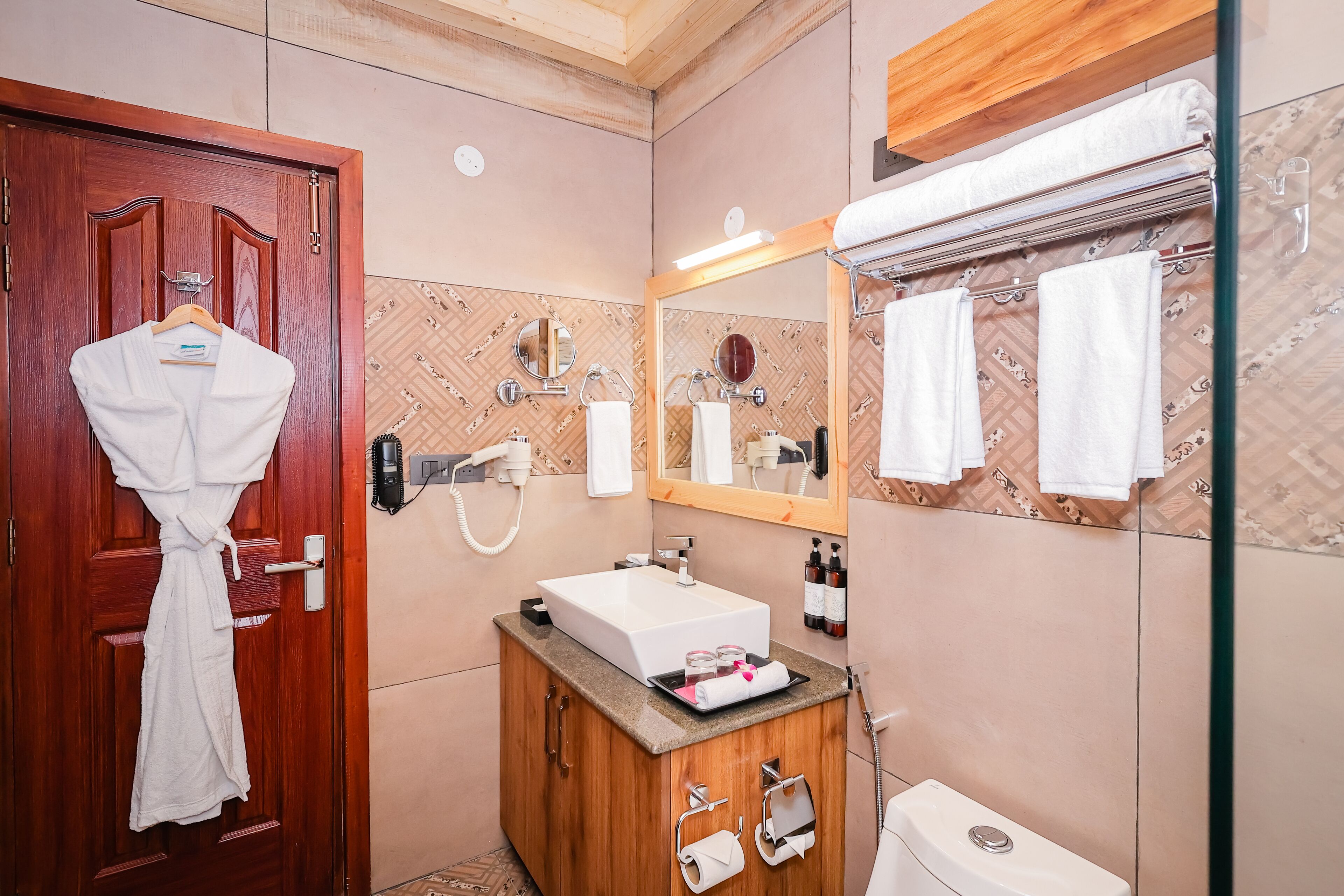 Un baño con puerta de madera, una bata blanca colgada en la puerta, un lavabo con espejo y un perchero con toallas.