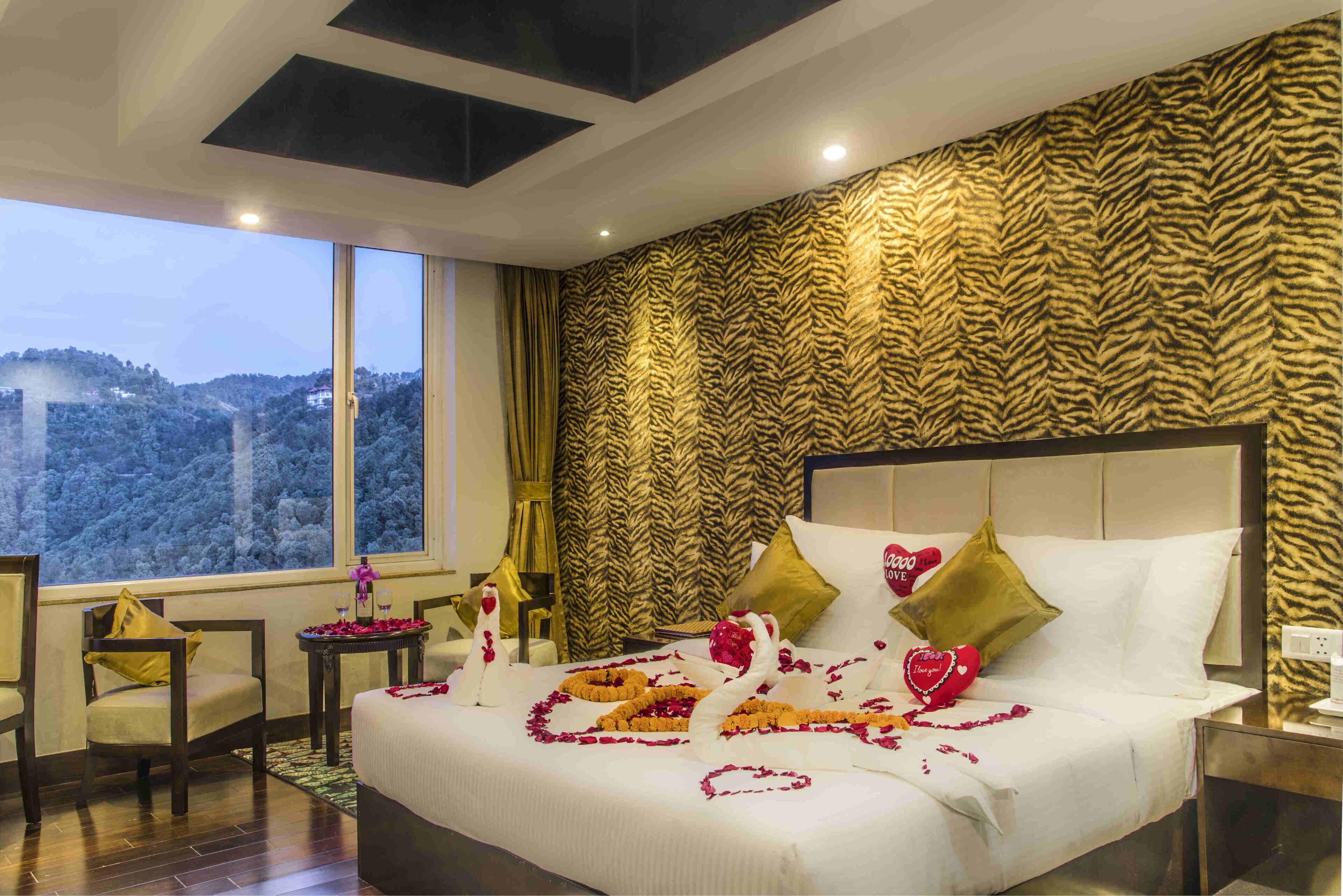 A hotel room with a bed adorned with rose petals and heart-shaped decorations, a large window offering a scenic view, and a tiger-print wall.