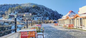 A snow-covered outdoor seating area with tables and chairs, overlooking a mountainous landscape with buildings. - Regenta Resort MARS Valley View (Shimla)