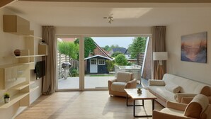 Een ruime woonkamer met een bank, salontafel en televisie. Er is een glazen schuifdeur naar een terras met buitenmeubilair en een kleine schuur.