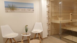 Een kamer met een schilderij van een strand, twee witte stoelen, een kleintje tafeltje met een plant en een houten kast met glazen deuren.