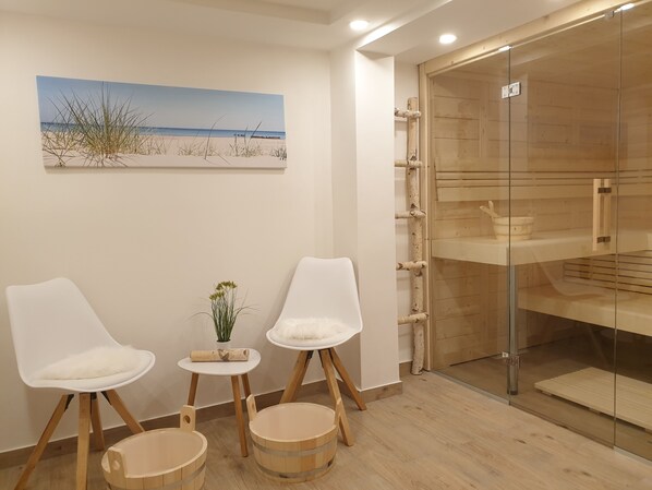Een kamer met een schilderij van een strand, twee witte stoelen, een kleintje tafeltje met een plant en een houten kast met glazen deuren.