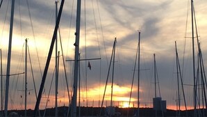 Een jachthaven met meerdere zeilboten afgemeerd, een zonsondergang op de achtergrond en een rustig wateroppervlak.