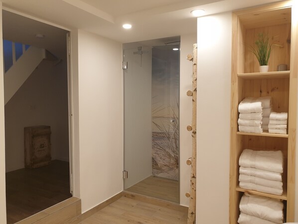 Une salle de bain moderne avec une douche à l’italienne en verre, une étagère en bois où sont pliés des serviettes, et un escalier apparent menant à une autre pièce.