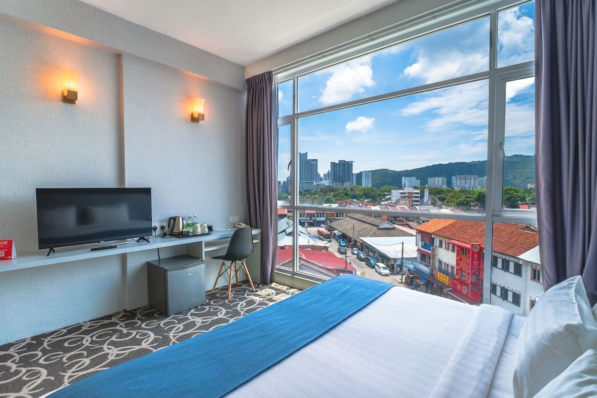 A hotel room with a bed, a desk with a TV, a chair, and a large window with a city view.