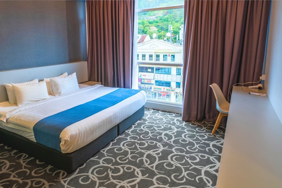 A hotel room with a bed, a window with curtains, a patterned carpet, and a chair.
