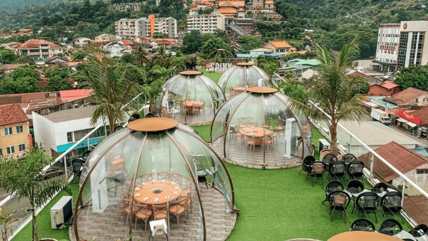 Eine Dachterrasse mit Kuppelartigen Konstruktionen aus Glas, Tischen und Stühlen, umgeben von Palmen und Gebäuden.
