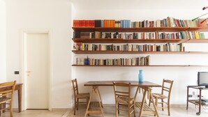Ein Zimmer mit einem Holzesstisch, vier Stühlen, einem Bücherregal mit mehreren Ablagen und einem Fernseher auf einem Schrank.