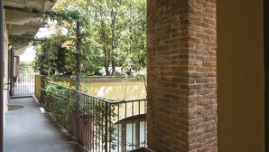 Un balcón techado con un pilar de ladrillo, barandilla metálica y vista a un patio con árboles y un edificio al fondo.